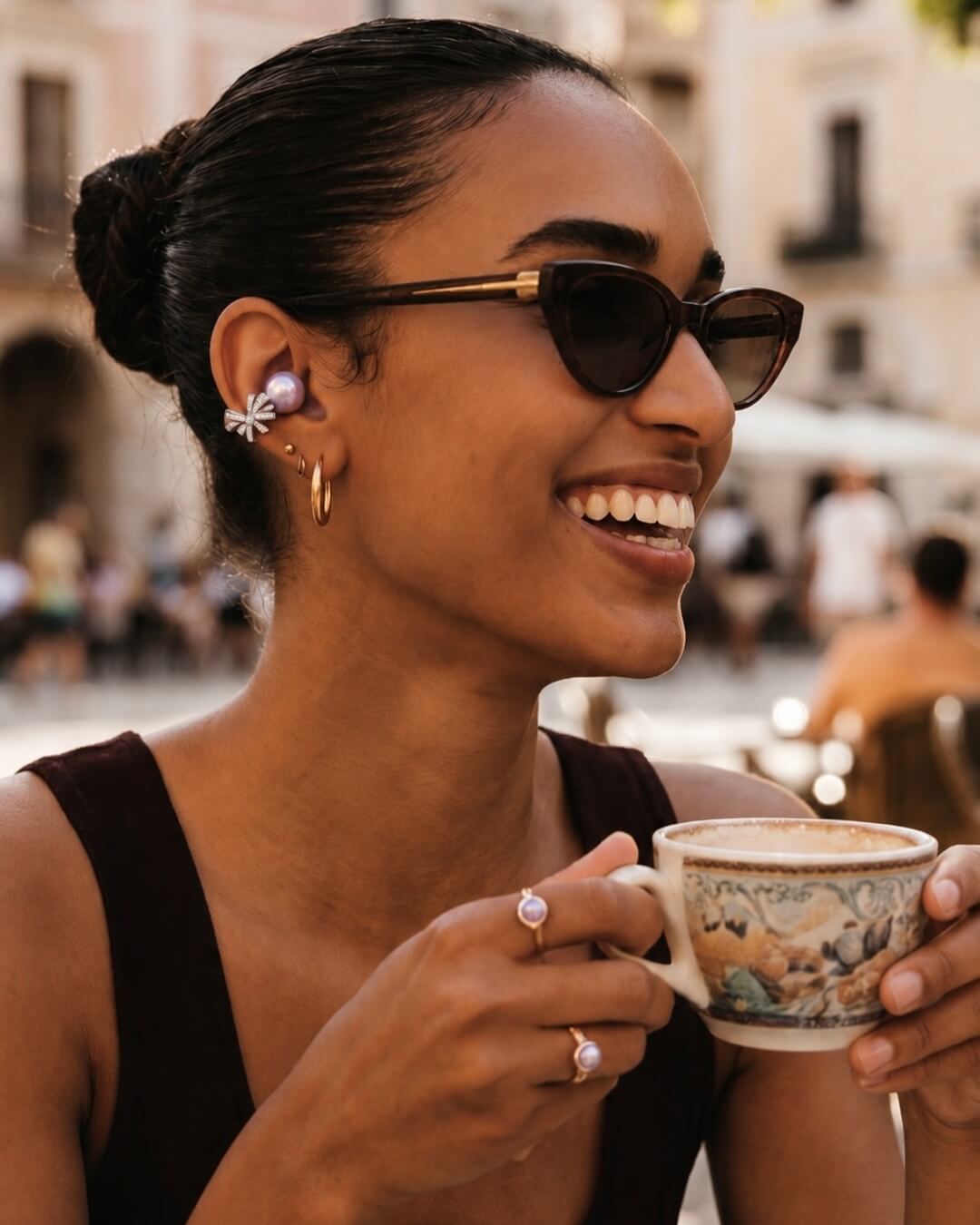 A smiling woman wearing sunglasses and a pearl ear cuff holds a small, ornate coffee cup outdoors.