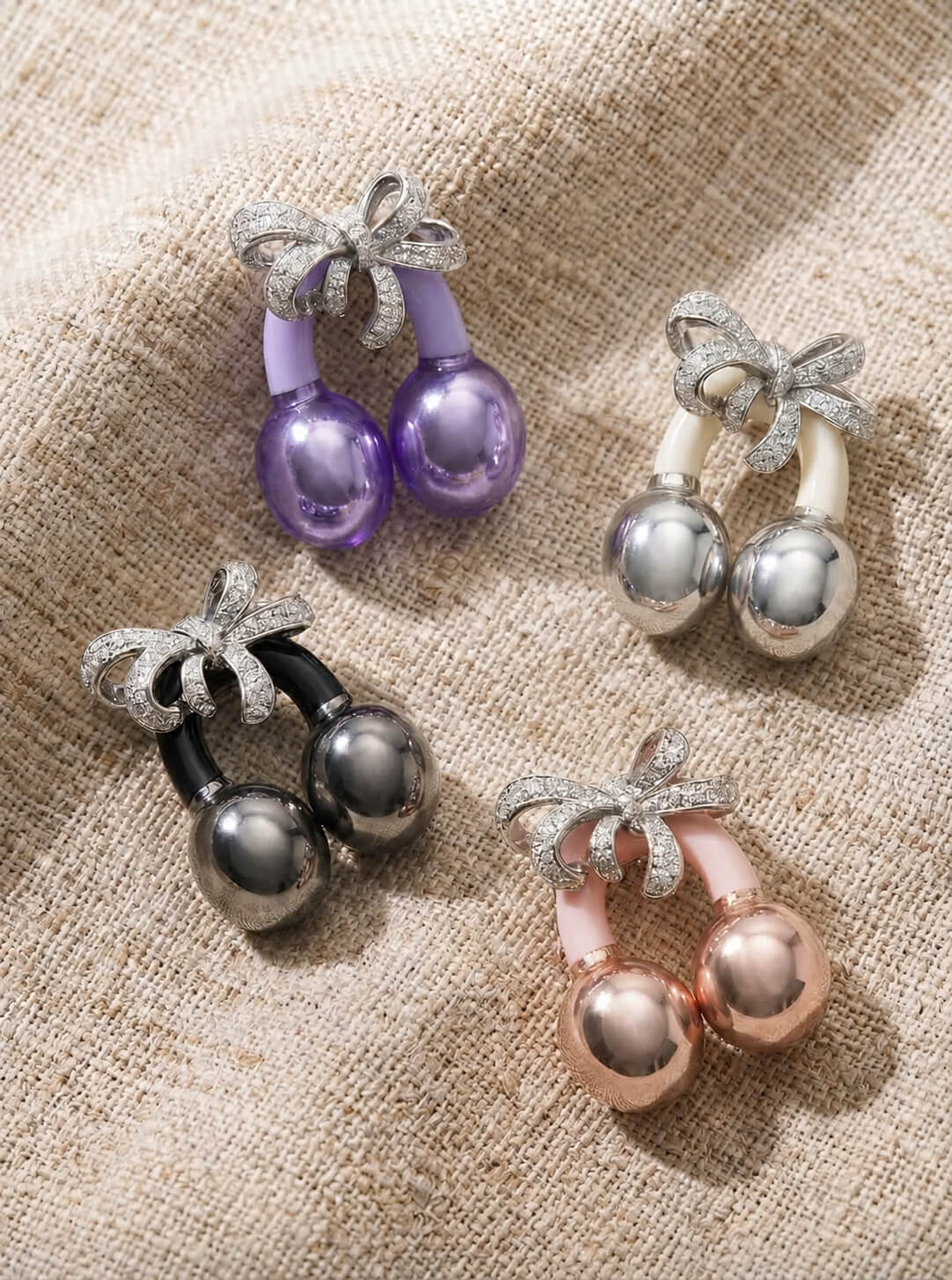 Four cherry-shaped brooches with diamond bows in various colors, resting on a beige woven fabric.