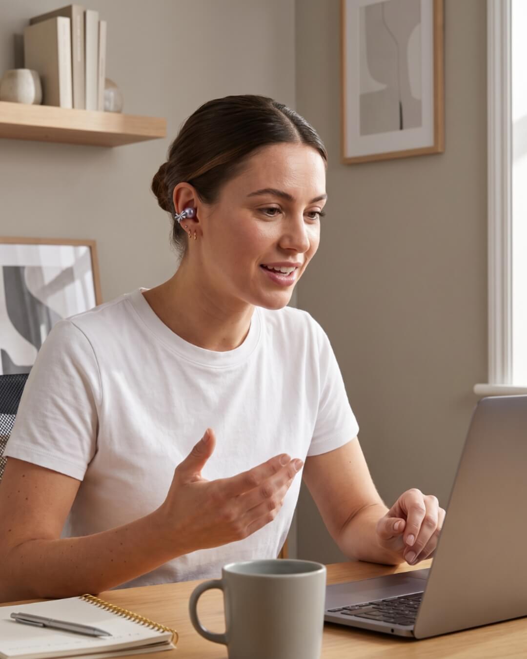 A woman wearing a hearing aid sits at a desk and gestures while on a video call.