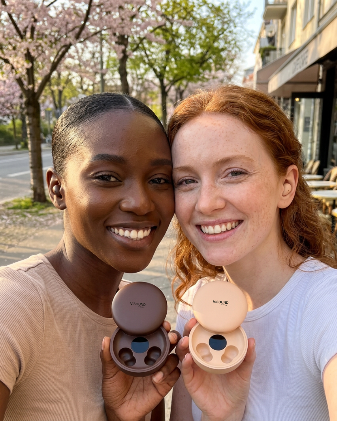 Two smiling women stand outside under blooming trees, each holding an open case of Visound wireless earbuds.