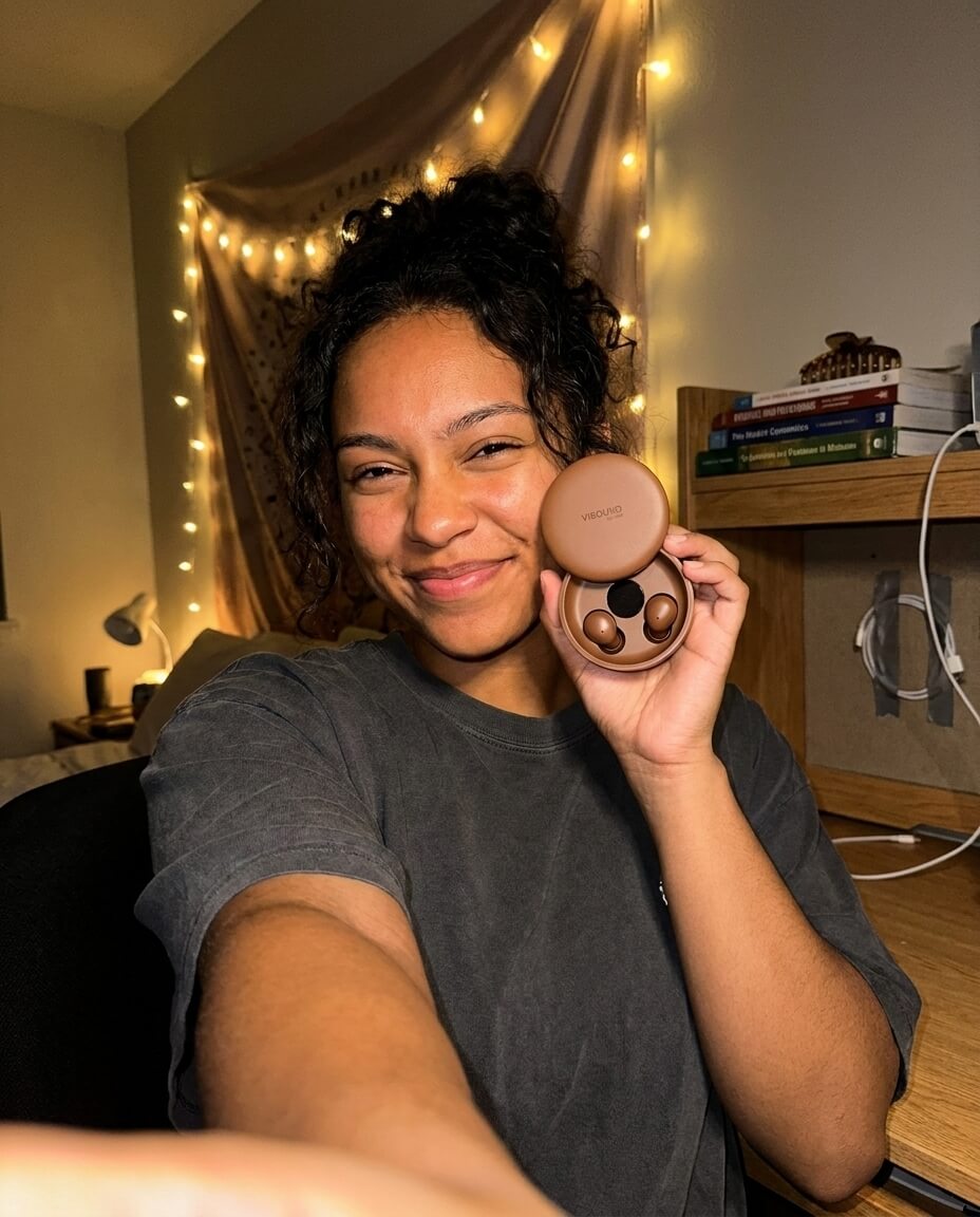 A smiling young woman takes a selfie in her room, holding an open case of brown wireless earbuds.