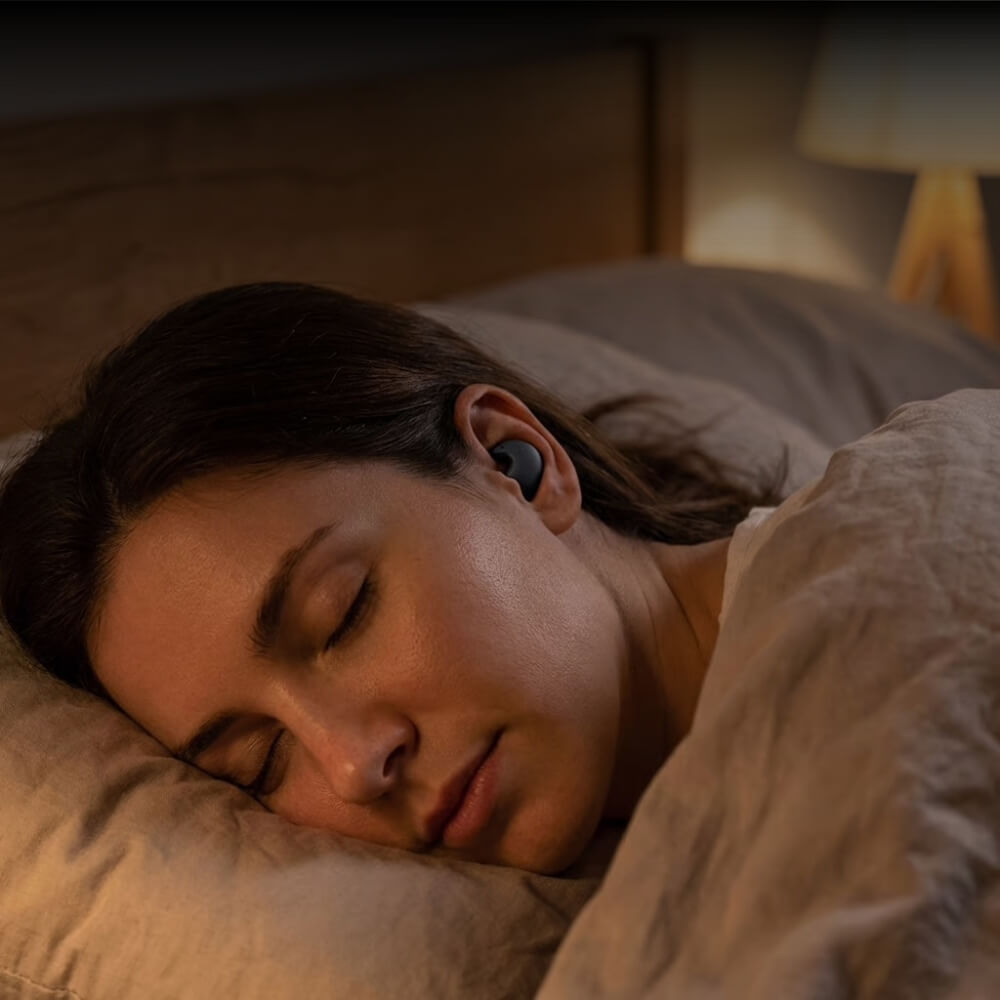A woman with her eyes closed sleeps in bed while wearing a black earbud.